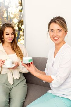 Beautician offering product for the young woman holding a white plastic jar with a cream sitting on the sofa