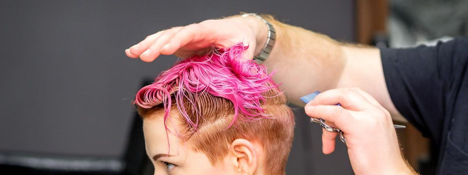 Haircut of dyed short pink wet hair of young caucasian woman by a male hairdresser in a barbershop