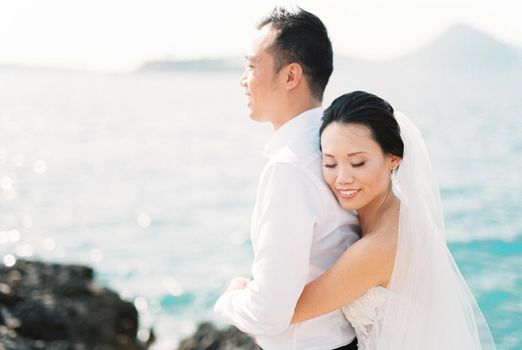 Bride in a veil hugs groom from behind on a rock by the sea. High quality photo