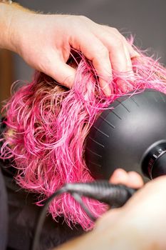 Drying short pink bob hairstyle of a young caucasian woman with a black hair dryer with the brush by hands of a male hairdresser in a hair salon, close up