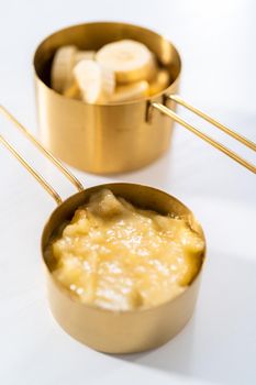 Mashed overripe bananas and sliced riped bananas in metal measuring cups.