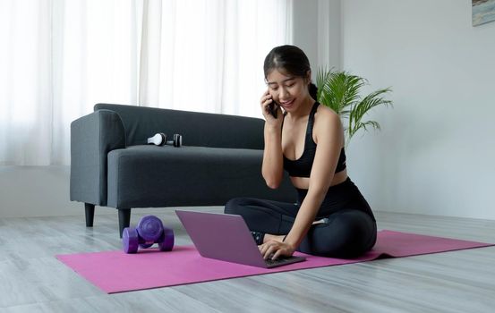 Asian sporty woman in sportswear working out and using laptop and call phone at home in living room, sitting on the floor with dumbbells on yoga mat. Sport and online training concept.