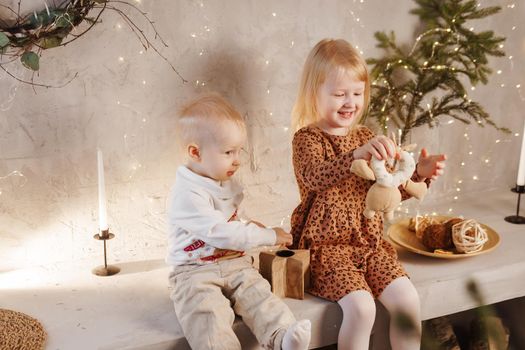 Little brother and sister play on Christmas eve in a beautiful house decorated for the New Year holidays. Children are playing with a Christmas gift. Scandinavian-style interior with live fir trees and a wooden staircase.