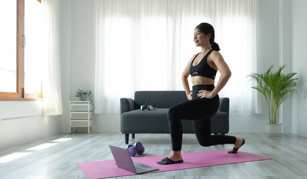 Smiling Asian woman doing yoga shoulder stretching online class from laptop at home in living room. Self isolation and workout at home..