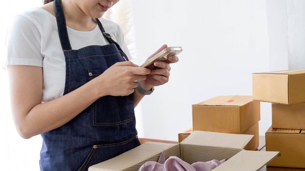 Hands of Asian woman entrepreneur holding smartphone with cardboard boxes, computer laptop, online selling equipments. Packing, business and technology concept. Top view, copy space.