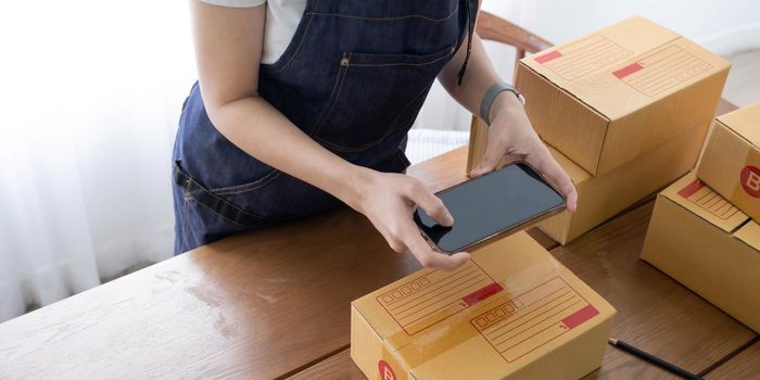 Hands of Asian woman entrepreneur holding smartphone with cardboard boxes, computer laptop, online selling equipments. Packing, business and technology concept. Top view, copy space.