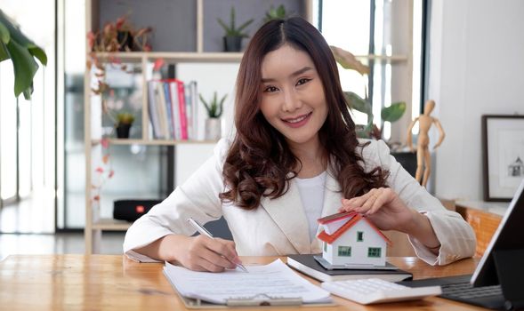 Miniature house in the hands of an Asian woman real estate agent home loan working at the office. Looking at the camera..