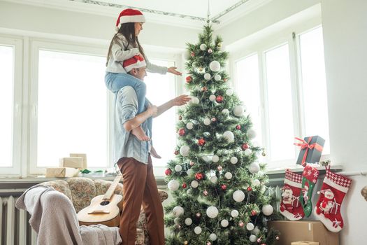 Family decorating a Christmas tree. Young man with his daughter on his shoulders helping her decorate the Christmas tree.