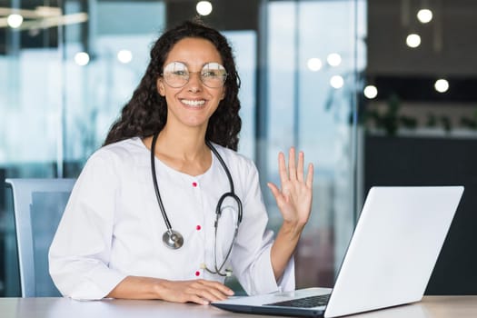 Portrait of a young Hispanic woman doctor sitting in a medical gown with a stethoscope in the office. Works with patients online with a laptop. He looks at the camera, smiles, waves his hand