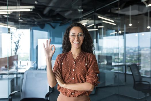 Portrait of a young beautiful hispanic woman freelancer, IT worker, SEO in glasses stands in the office, looks at the camera, smiles, shows ok with her hand.