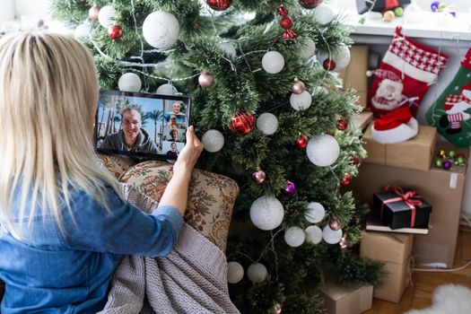 A middle-aged blonde woman uses a tablet for a video call while sitting in an armchair.