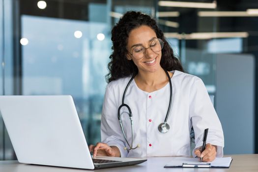 Online training. Young beautiful Latin American girl student doctor studies, takes exam online remotely at home from laptop. Sitting at the table in a medical gown, writing in a notebook.