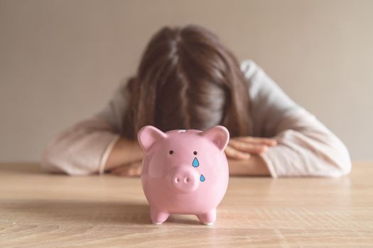 Sad girl leaned on the table, covering her face with hands, in the foreground a crying piggy bank with painted tears. Woman lost her savings, was fired, and is experiencing financial difficulties.