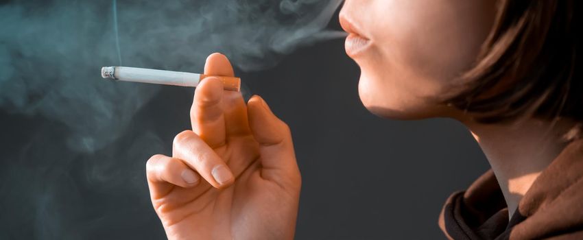 A young beauty girl is smoking, a woman is stressed and holds a cigarette in her hands, staying near the window of her apartment, close-up view.