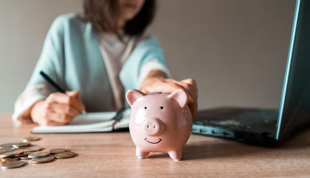 A young girl makes calculations, checks her finances, makes notes in a notebook and sets aside some of the funds, savings in a piggy bank for personal expenses and future development.