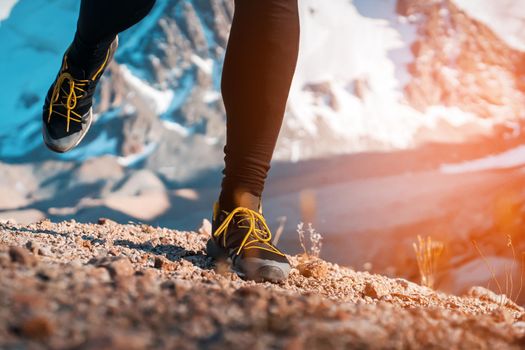 Athlete runner legs run on a trail along a rocky path close-up. Fitness and warm-up, young man with snow-covered mountain peaks in the background and open space around him at sunset.