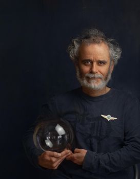 a colorful man with a curled beard and a silvery mustache holds a transparent glass ball in his hand and looks forward smiling a little on his chest civil aviation badge High quality photo