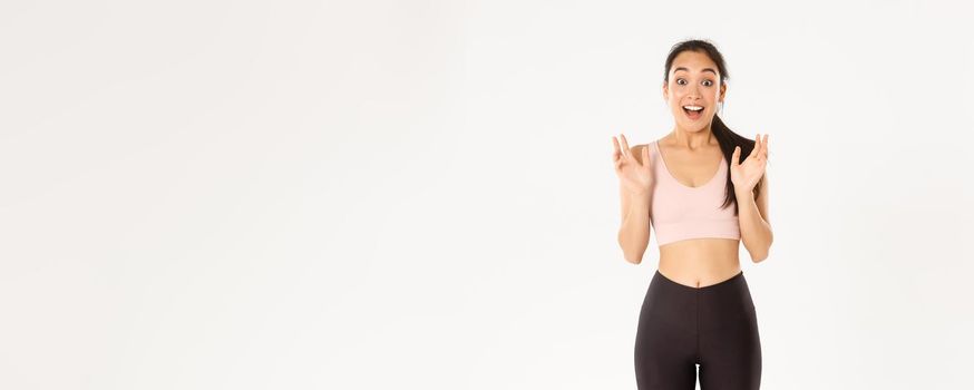 Sport, wellbeing and active lifestyle concept. Happy enthusiastic fitness girl, asian sportswoman clasp hands from fantastic news, gasping amazed, standing over white background.