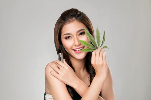 Portrait of young ardent woman with healthy fresh skin holding green hemp leaf and cbd oil. Combination of beauty and cannabis concept.