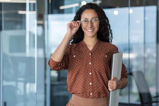 Portrait of a young Hispanic woman. Latin American woman architect and designer stands in the office with projects and drawings in her hands, holds glasses in her hand, looks at the camera, smiles.