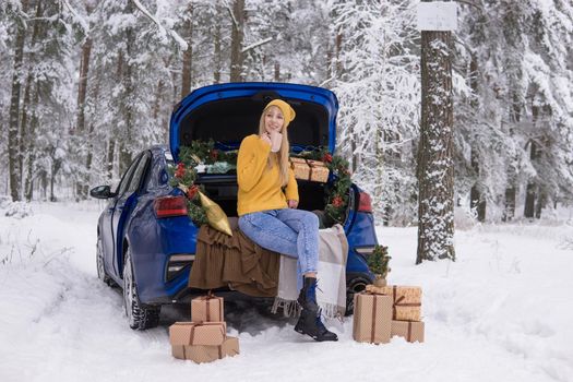 A woman in a winter snow-covered forest in the trunk of a blue car decorated with Christmas decor. The concept of Christmas and winter holidays.