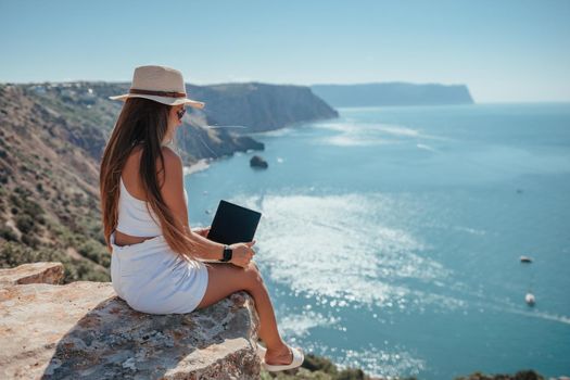 Successful business woman in yellow hat working on laptop by the sea. Pretty lady typing on computer at summer day outdoors. Freelance, travel and holidays concept.