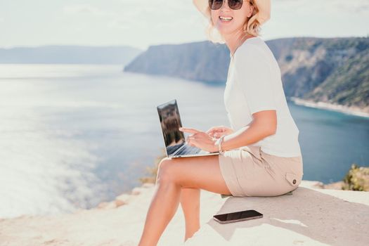 Successful business woman in yellow hat working on laptop by the sea. Pretty lady typing on computer at summer day outdoors. Freelance, travel and holidays concept.