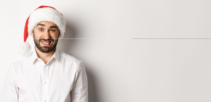 Close-up of awkward guy in santa hat apologizing, feeling uncomfortable, standing over white background. Christmas concept.