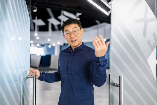 Asian businessman boss invites to his team, man looks at camera and waves, manager in glasses and shirt works inside modern office building.