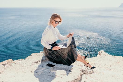 Successful business woman in yellow hat working on laptop by the sea. Pretty lady typing on computer at summer day outdoors. Freelance, travel and holidays concept.