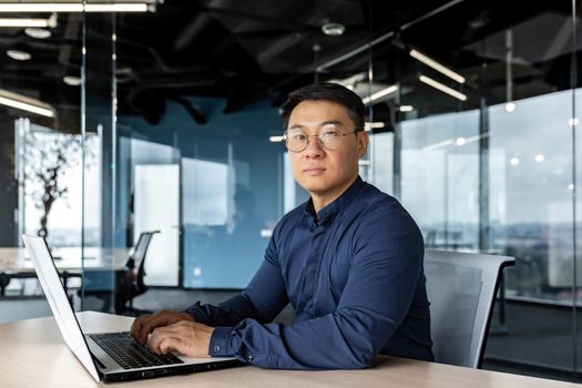 Serious and focused businessman looking at camera, asian man working inside modern building, man wearing glasses and casual shirt using laptop.