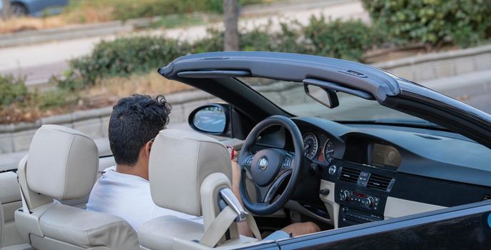 A man sitting inside in his convertible car waiting for someone while looking his mobile phone. Business and leisure concept