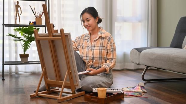 Pleasant female artist sitting on floor in bright living room and painting with watercolor on canvas. Art and leisure activity concept.