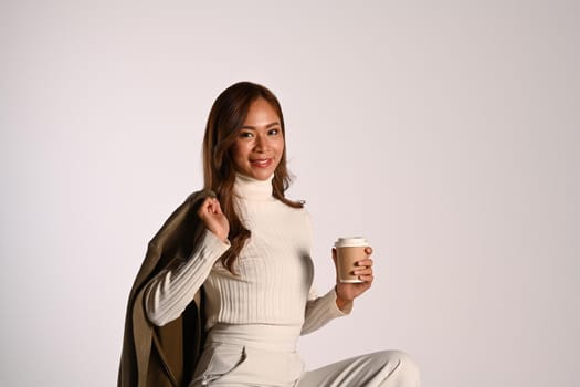 Portrait of stylish young woman in trendy clothes holding paper cup sitting over white background. Fashion, beauty concept.