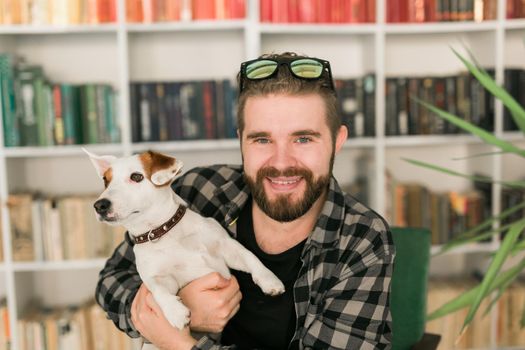 Happy male owner of jack russell terrier dog, feels responsibility of caring about pet, standing against bookshelves background. People and relationship with animals.