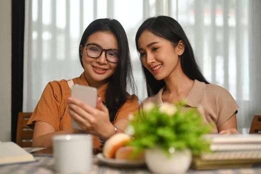 Two happy asian female students resting after reading book and checking social media on mobile phone.