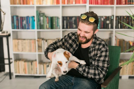 Happy male owner of jack russell terrier dog, feels responsibility of caring about pet, standing against bookshelves background. People and relationship with animals.