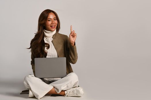 Smiling young woman in trendy clothes sitting with laptop and pointing at copy space over white studio. People and technology concept.