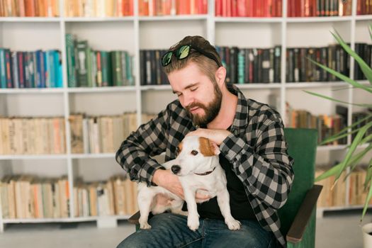 Happy male owner of jack russell terrier dog, feels responsibility of caring about pet, standing against bookshelves background. People and relationship with animals.