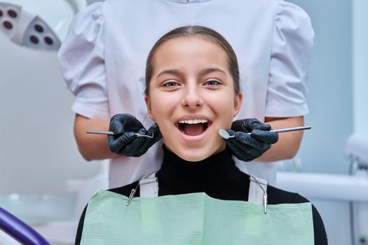 Portrait of young teenage girl in dental chair with hands of doctor with tools. Female teenager smiling with teeth looking at camera in dentist office. Adolescence hygiene treatment dental health care