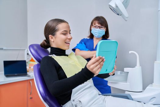 Happy young teenage female with mirror sitting in dentist chair smiling looking at healthy teeth, with dentist doctor in office. Dentistry, hygiene, treatment, dental health care concept