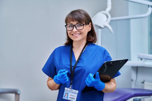 Portrait of female dentist in office. Middle aged doctor nurse looking at camera with professional badge clipboard near dental chair. Dentistry, medicine, specialist, career, dental health care concept