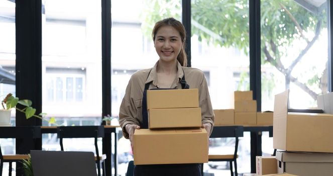 Young attractive asian woman owner startup business look at camera work happy with box at home prepare parcel delivery in sme supply chain, procurement