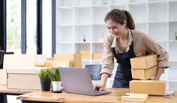 Young smiling beautiful owner asian woman freelancer sme business online shopping working on laptop computer with parcel box at home - SME business online and delivery concept.