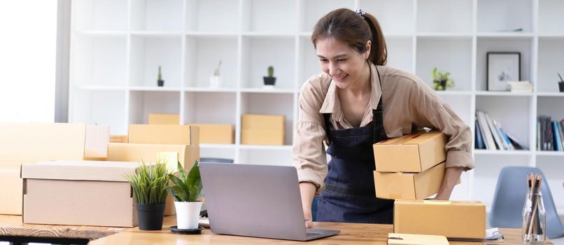 Young smiling beautiful owner asian woman freelancer sme business online shopping working on laptop computer with parcel box at home - SME business online and delivery concept.