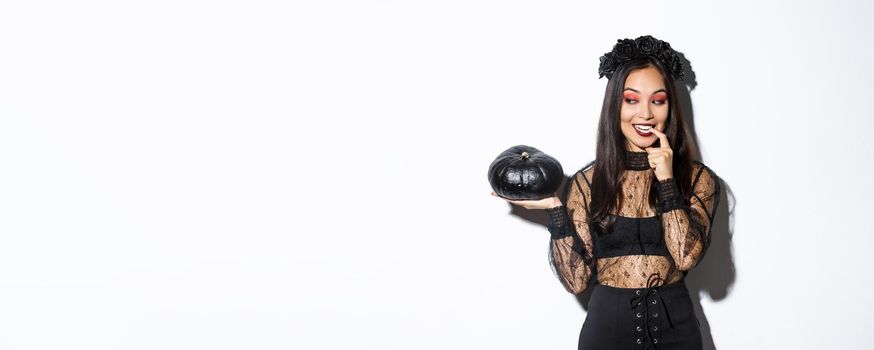 Image of sexy asian woman in witch costume looking at black pumpking and smiling, standing over white background.