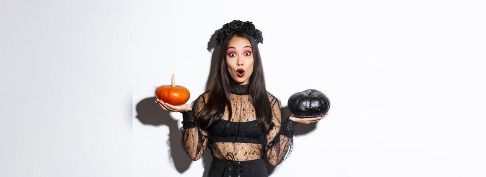 Portrait of surprised asian girl in witch costume holding two pumpkins and gasping amazed while looking at camera, standing over white background.