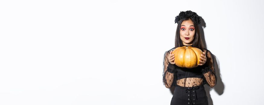 Portrait of excited pretty asian woman in halloween costume, dressed-up as witch or widow and holding pumpkin, standing over white background.