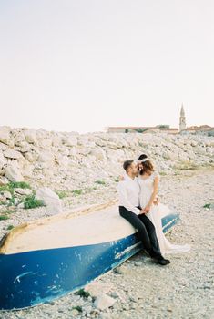 Bride stands next to groom sitting on an upside down fishing boat on the shore. High quality photo