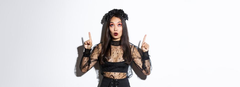 Surprised attractive asian woman in witch outfit pointing fingers up, showing halloween banner, promo of party. Beautiful female in black gothic dress looking amazed.